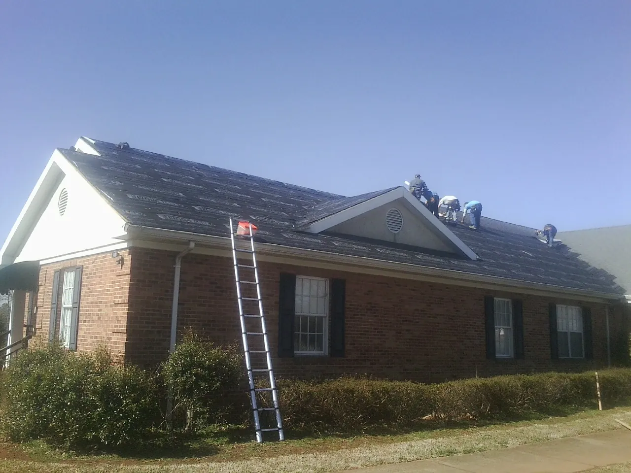 Crew tearing shingles off a brick ranch roof with a ladder set — Metal Roof Repair work by our crew in Williams